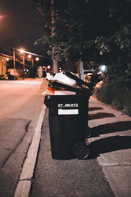 A black wheeled rubbish bin with the label 'St. John's' is positioned on a concrete sidewalk next to a street curb at night. The lid is open, revealing a mix of waste materials including cardboard box