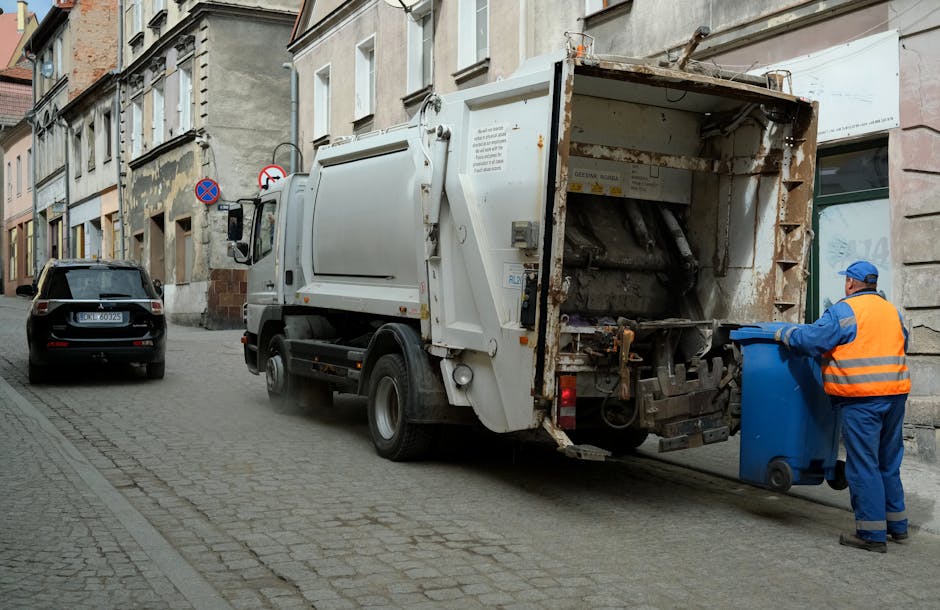 A man with dark hair, dressed in a black T-shirt and dark trousers, is bent over and depositing waste into a modern, stainless steel trash bin located along a paved outdoor walkway. He holds a large w