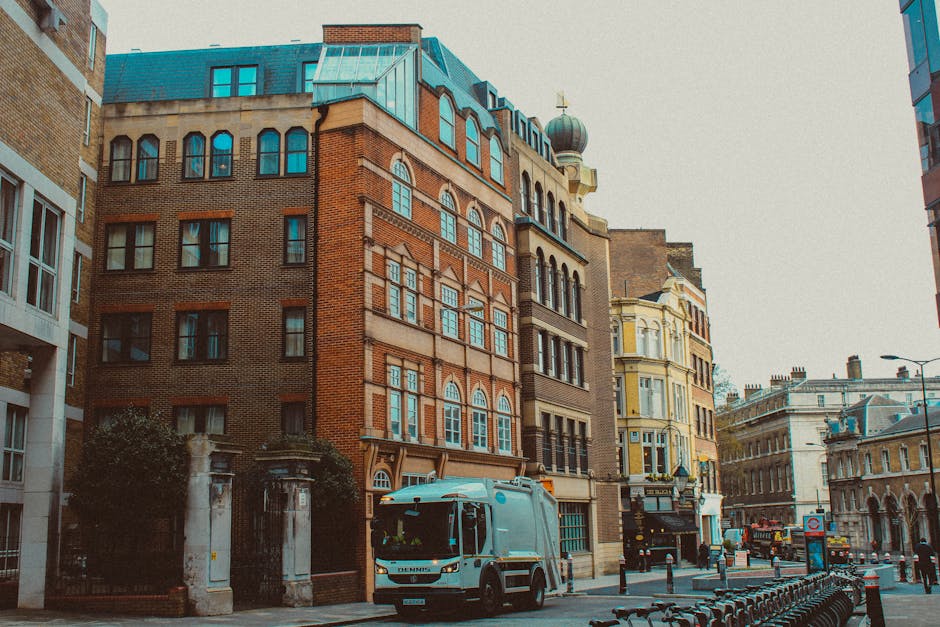 A street scene in an urban area featuring a row of historic multi-storey buildings with various architectural styles. The central building is constructed with reddish-brown brick and has large rectang